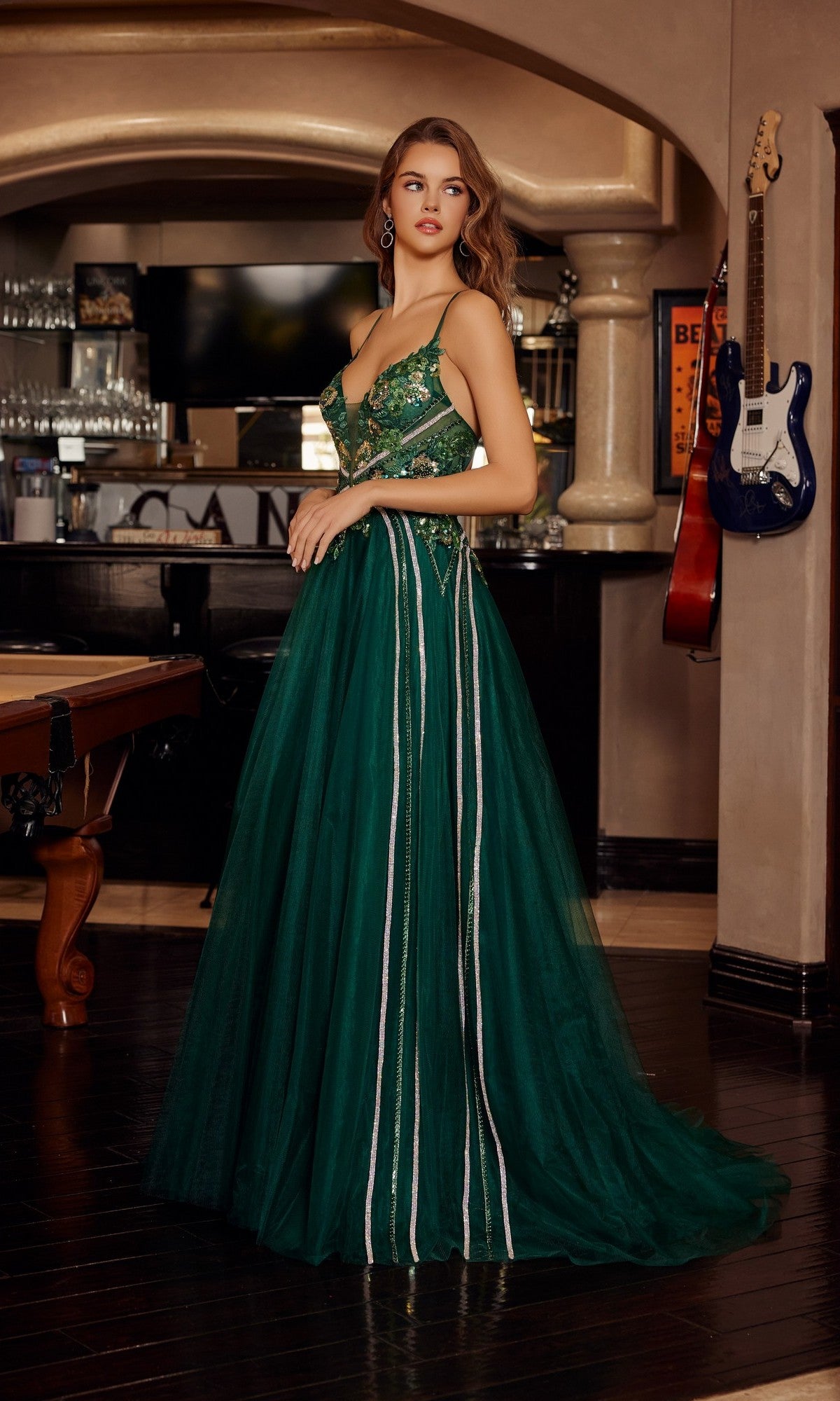 A woman stands indoors wearing the Nox Anabel G1795, a long, dark green prom dress with sequins and thin straps. In the background are a pool table and guitars.